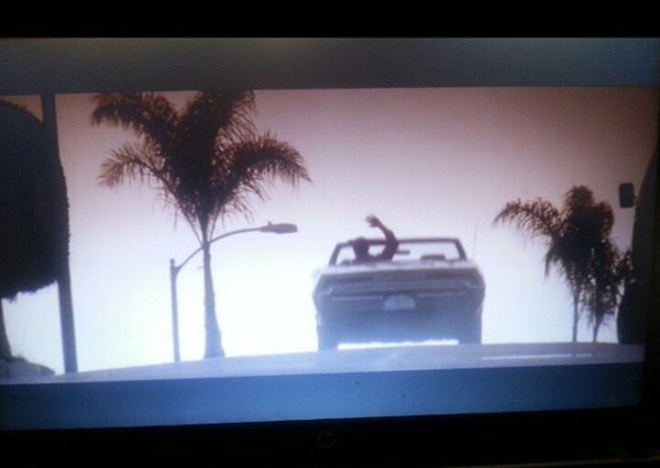 Screen shot a topless convertible car driving up a street lined with me Palm trees. The driver looks like he is waving goodby. Hasta la vista.